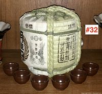 Barrel flask wrapped in woven straw with Japanese writing, surrounded by six small brown saki cups.