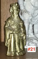 Photo of brass or bronze Chinese figure showing front view with long beard, traditional robes, and headdress, placed on wooden shelf.