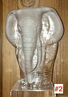 Front view of clear engraved glass elephant head sculpture showing detailed ears, trunk and tusks on rectangular base.