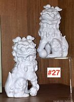 Pair of white glazed Asian porcelain foo dogs, one with paw on a ball, displayed side by side on wooden background.