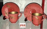 Pair of Asian lacquerware demi-tasse cups and matching saucers, coral-red with gold bird designs, displayed on stands.