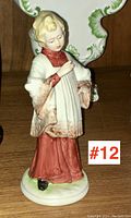 Porcelain figurine of a child wearing a red skirt and white cloak with brown detailed trim, shown from the front.