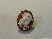 Front view of oval shell cameo depicting profile of a young woman with flowing hair in white relief against brown background, set in gold prong setting.