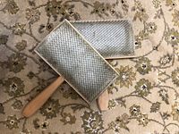 Two fleece carders with wooden handles and dense metal teeth placed on a patterned carpet seen from front side.