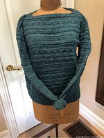 Front view of the hand-knit teal sweater on a dress form showing textured knit patterns and long sleeves.