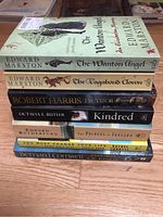 Photo showing a stack of 9 paperback books with historical and fictional themes on a wooden floor