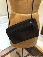 Front and side view of black handbag with crossbody strap worn on dress form, showing exterior zip pocket and texture.