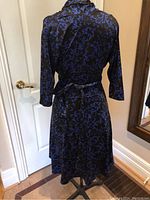 Back view of black dress with blue floral/vine pattern, 3/4 length sleeves, and tie belt around waist on dress form.