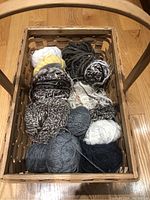 Top-down image of a rectangular woven basket containing various yarns in neutral tones including white, gray, black, cream, and a small amount of yellow. The yarns vary in thickness and type including hand spun and regular yarns.