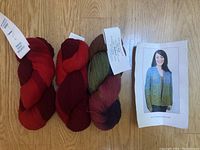 Three colorful skeins of yarn laid on wooden surface with labels visible, showing red, green, brown, purple, and pink tones.