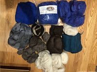 Overview of various colored roving and batt fibers arranged on a wooden floor including blue, grey, black, white, brown, and teal fibers.