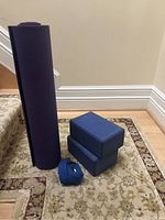 Extra long rolled purple yoga mat standing upright with two blue yoga blocks stacked together next to it. A blue yoga strap is coiled on the ground beside the blocks.