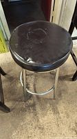 Photo showing black vinyl padded round bar stool seat with chrome metal legs and circular footrest ring on concrete floor.