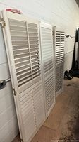 Two white louvered doors standing against a white wall with hinges attached. Shows overall height and slatted design.