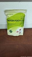 Photo of a sealed, green and white resealable bag marked 'Detox Foot Patch' containing 10 patches, placed on a wooden surface against a white wall.