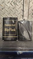 Photo of the stainless steel hip flask next to its original black packaging box with gold lettering.