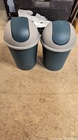 Front view of two small garbage bins with turquoise bases and light grey swing-top lids placed on a wooden floor.