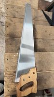 Full view of a hand saw with wood handle laid flat on wooden surface, showing the metal blade and wooden handle details.