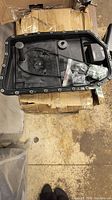 Black BMW transmission oil pan resting on a cardboard box, includes a sensor/tube and mounting hardware in plastic bags.