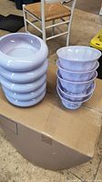Photo showing 11 stacked lilac ceramic bowls with scalloped edges and 5 stacked matching lilac colored rounded planter bottoms on a cardboard box.