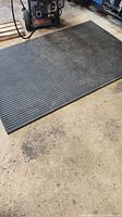 Black ribbed rubber mat lying flat on a concrete floor next to a piece of machinery