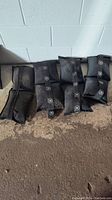 Four black sandbags lying on the ground against a white cinderblock wall. Each bag has a circular logo with text.