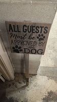 Wooden yard sign with text 'All Guests Must Be Approved By The Dog' featuring paw prints and attached to a wooden stake.