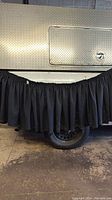 Black gathered fabric table skirt hanging on side of a vehicle, showing texture and length relative to a tire. Suitable for table edges, gathered styling.
