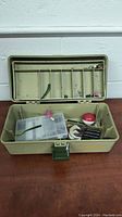 Open green fishing tackle box showing compartments with assorted fishing lures, hooks, bobbers, and a small plastic compartment box.