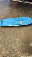 Blue lightweight plastic toboggan with contoured seat and yellow handles on a concrete floor