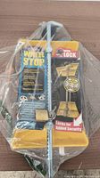 Photo of sealed metal wheel stop in plastic packaging with included brass padlock and keys visible. Packaging shows branding and product info with emphasis on lock security.