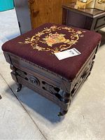 Full view of antique foot stool showing dark carved wooden base with intricate design, four turned legs with casters, and burgundy needlepoint cushion featuring a floral rose bouquet in the center.