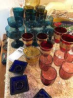 Overview of various teal blue goblets, red rimmed goblets, square blue candle holders and smaller glasses on countertop.