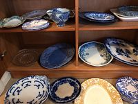 Wooden shelf displaying a variety of ceramic and porcelain serving dishes and plates in blue and white, including platters and small plates.