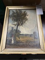 Framed oil painting showing a woman in white dress with red headscarf by a large tree in a rural landscape.