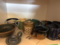 Shelf display of various pottery items: mugs, bowls, a garlic keeper, and candle holders in earth/blue color tones.