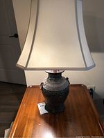 Full view of a decorative table lamp with a white paneled lampshade and ceramic urn-shaped base decorated with floral patterns and metal ring handles, sitting on a wood table.