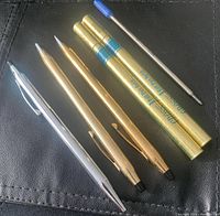 Three Cross writing instruments: two gold tone (one pen and one pencil) and one chrome pen, accompanied by three unopened ball pen refills in original packaging, plus one loose refill visible.