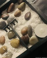 Close-up of zen garden tray with white sand, polished decorative stones in varying colors, small black rake, and white tealight candle