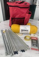 Red fabric knitting bag with assorted knitting needles, yellow Bernat Premium yarn, measuring tape, and boxed circular needles on tabletop.