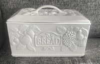 Front view of white ceramic breadbox showing raised botanical designs and embossed 'BREAD' label.