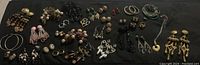 Wide view of many pairs of costume earrings arranged on dark cloth surface, showing variety in size, style and color
