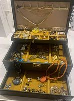 Open black faux leather three-tier jewelry box showing top hanging hooks with necklaces, plus middle and bottom trays holding assorted brooches, rings and necklaces