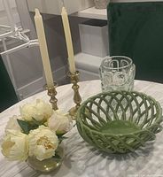Photo showing green lattice bowl, faux floral arrangement, brass candlesticks with taper candles, and heavy crystal vase all placed on a white marble table.