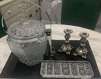 Photo showing vintage pressed glass lidded ice bucket with metal handle and rim, silver tone ornate candlesticks, pressed glass dish, and tongs on a table.