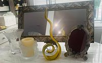 Photo showing whole lot items on table: rectangular silver decorative framed mirror, yellow art glass sculpture, silver ornate photo frame holding 5x7 photo, clear etched glass vase, frosted angel candleholder.