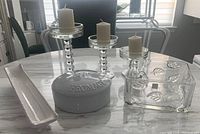 Photo showing all items on the table: a white ceramic lidded cheese keeper with 'FROMAGE' embossed on the lid, a square clear glass bowl with leaf patterns, a long white cracker dish, and three clear glass candle holders of different heights with pillar candles on top.