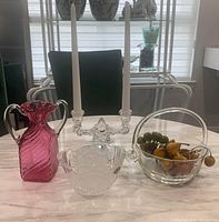 Photo of cranberry glass vase with two handles, crystal serving basket with handle, double candleholder with white taper candles, and crystal dish containing artificial fruit. Items shown on a marble table in natural light.