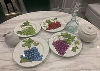 Photo showing four ceramic fruit plates with grape designs in green, red, purple, and blue colors, white pear-shaped lidded dish, white lidded sugar bowl, and glass decanter with grape stopper on round marble table.