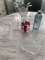 View of the glass items on a table with a water bottle for size reference. Includes a tall fluted glass candleholder, 2 smaller flower-shaped candleholders, glass Santa plate, and square glass gift box wrapped like a present.